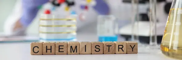 Word Chemistry made of wooden cubes near laboratory glassware on table. Blocks with letters against blurry scientist with molecule model in hands