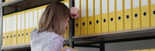Exhausted woman fails finding right materials from archive for professional research. Secretary leans on rack with yellow ring binders on shelves