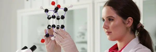Laboratory worker in white coat examines model of molecule. Positive woman works with chemical reagents in laboratory on blurred background side view