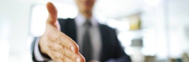 Takım elbiseli adam el sıkışmak için elini uzatıyor. Erkek ofis çalışanı, bulanık arka planda oturan meslektaşını ya da müvekkilini selamlıyor