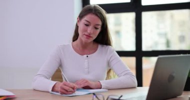 Odaklanmış, gülümseyen genç bir kadın dizüstü bilgisayarın yanındaki masaya oturur ve not defterine bilgi yazar. Önemli notlar al ve kişisel notları kaydet.