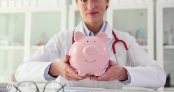 クリニックでピンクのピギーバンクを持っている女性医師の看護師 病院予算 診療費について — ストック動画