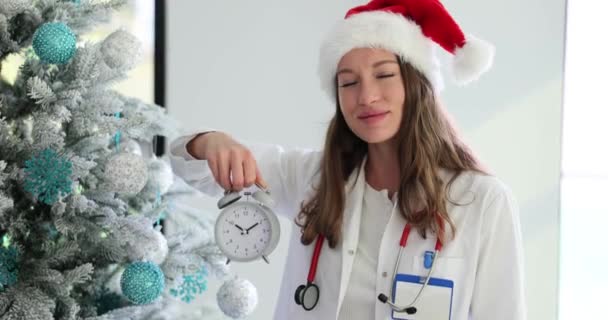 女性医師はクリスマスツリーの背景に対して目覚まし時計を保持します クリスマス休暇や新年中に医療援助をタイムリーに求める — ストック動画