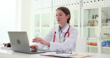Laptop klavyesinde daktilo eden, klinikteki iş yerinde oturup kapatan bir kadın doktor. Tıp çalışanı bilgisayarla çalışır ve metin yazar, uzaktan çalışmayı bitirir ve masada oturur