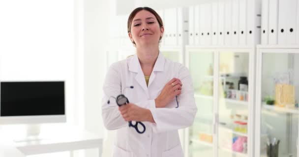 快乐的年轻女医生穿着白色的医疗外套 手持听诊器 看着相机 在医院办公室摆出一副自信的女医生的样子 — 图库视频影像