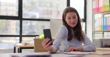 Genç kadın sekreter şirket ofisinde cep telefonuyla poz değiştirirken selfie çekiyor. İş yerinde tembellik ve modern teknolojiyle anı yakalamak