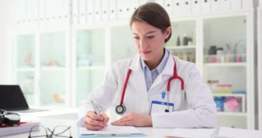 Odaklanmış ifadesi olan kadın doktor, hasta bilgilerini masa başında oturarak hafif bir önerme ile dolduruyor. Klinikte hasta için rapor doldurma ve tedavi