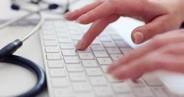 Doktor beyaz klavyeye hasta tanı bilgilerini giriyor. Stetoskop, klinikteki ağır çekimde hafif klavyenin yanındaki ofis masasında.