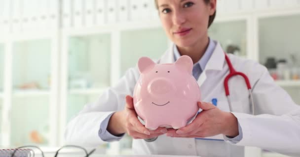 Ärztin Mit Stethoskop Zeigt Rosafarbenes Sparschwein Mit Ersparnissen Drinnen Schreibtisch — Stockvideo