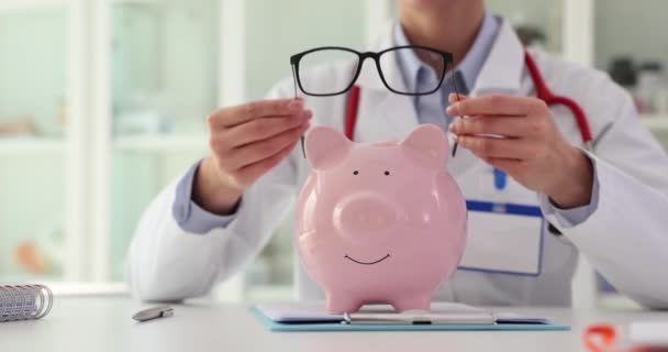 女性医師は 病院のオフィスのテーブルの上にピギーバンクで笑顔の眼鏡をかけます 病気の患者の治療のためにお金を集める慈善団体 キャッシュセーブメント — ストック動画