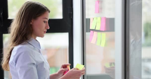 女性のマネージャーはオフィスの透明なガラス壁にノート紙を貼ります カラフルなステッカーと中小企業の開発を持つビジネスマン — ストック動画