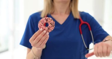 Kadın doktor donut tutuyor ve klinikte baş parmağını sallıyor. Tatlıdan ve diyetten kaçınma