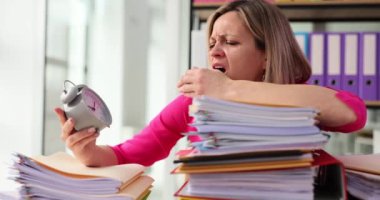 Çalar saatin kapanması ve yorgun kadın işçinin belgelerle dolu bir yığın klasörün üzerinde uyuması. Fazla çalışma ve fazla mesai