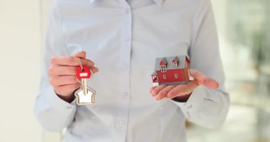 Kadın emlakçı, iki katlı bina ve anahtarın küçük modelini elinde tutan müşteriye ev seçeneklerini sunuyor. Emlak alım ve kiralama ajansı