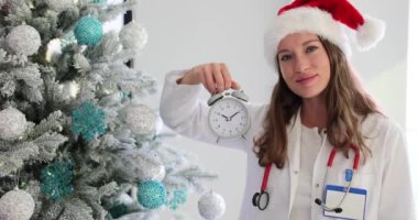 Gülümseyen kadın doktor, Noel ağacının yakınındaki bayramı kutlamak için alarmlı saati işaret ediyor. Beklenti ve kutlama ağır çekim