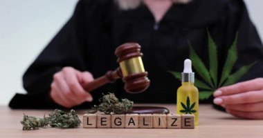 Sözcük, yargıç önünde tokmakla tahta küplerden yapılmış. Kadın şişenin yanında taze marihuana yaprağı tutuyor.