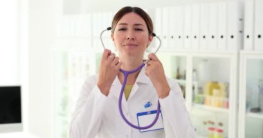 Kadın doktor, klinikteki hastanın sağlığını kontrol etmek için stetoskop takıyor. İş üniformalı sıhhiyeci ağır çekimde çalışacak ekipmanlarla ofiste duruyor.