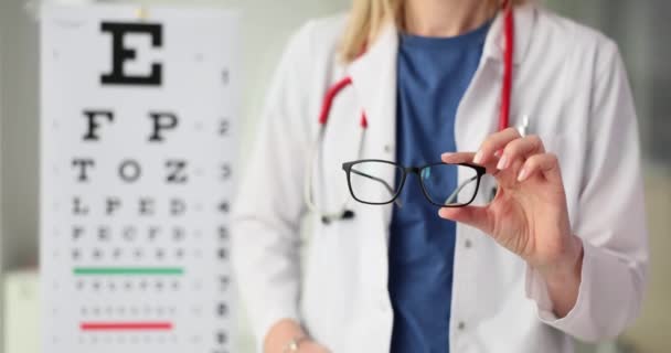Oculistは 視力の遅い動きのクライアントの能力を調べた後 患者の矯正眼鏡を提供しています ドクターはテストSnellenチャートでメガネを保持します — ストック動画