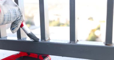 İş eldiveni teslim eden adam, balkon konseptinde gri boyalı fırçayla paslı metal boru boyuyor.