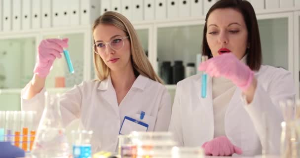 女性の化学者と研修生の実験助手は 化学実験室で青い液体を有する試験管を調べる 研究成果コンセプトの比較 — ストック動画