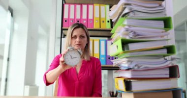 Kadın kağıt klasörlerle dolu bir masada çalar saatle elini uzatıyor. Muhasebeci, öğle saatlerinde iş yerinde çalışmaktan bıktığını gösteriyor.
