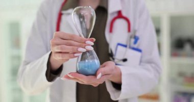 Kadın doktor muayenehanede kum saatini ters çeviriyor. Terapist, tedavi prosedürünü antika aletlerle yavaş çekimle ölçüyor