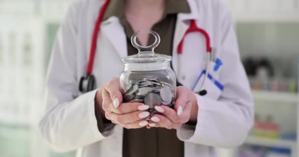 女性医師は クリニックオフィスにコインを持ったガラス瓶を持っています 医療従事者は 病院の医療コンセプトのための予算を実行します 治療のための支払い — ストック動画