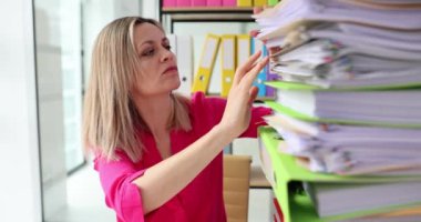 Kadın iş çalışanı, bitirilmemiş arşivleri aramak ve kontrol etmek için el ile kağıt yığınları ile çalışıyor. İşyerindeki yoğun masadaki kağıt dizinlerindeki belgeler