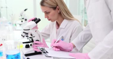 Mikroskop kullanan bilim adamı maddeyi inceler ve meslektaşı laboratuardaki not defterine test sonuçlarını yazar. Bilimsel Tıbbi Kimya Merkezi 'ndeki meslektaşların takım çalışması