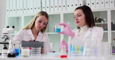 Laboratuvar asistanı ve bilim adamı rapora not alır ve mavi sıvıyla şişeyi tutar. Bilim adamı, laboratuardaki meslektaşıyla birlikte gözlemler ve sonuçlar yazar.