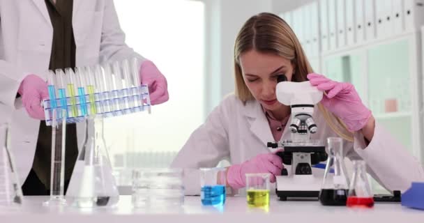 Wetenschapper Kijkt Naar Microscoop Collega Plaatst Reageerbuizen Met Gekleurde Vloeistoffen — Stockvideo
