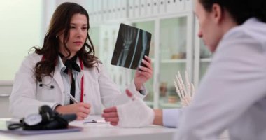 Yoğunlaşmış kadın doktor, ellerinin röntgenini inceliyor. Hastanın yanında bandajlı ellerle notlar tutuyor. Hastanedeki uzmanla randevu.