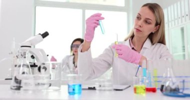 Ciddi kadın teknisyen test tüplerindeki renk sıvılarını laboratuvarda yavaş çekimde karşılaştırıyor. Bilimadamı tıp laboratuarındaki reaktörlerle numuneleri araştırıyor.