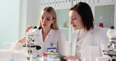 Kadın bilim adamı mikroskoba bakar ve kimya laboratuvarında çalışan bir meslektaşı işaret eder. Kadınlar numunelerle başarılı bir deney yapıyorlar.