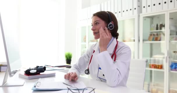 ポジティブな女性医師は 病院のオフィスでヘッドフォンとコンピュータを通じて患者とコミュニケーションをとります クリニックでのオンライン予約でクライアントとのコミュニケーション — ストック動画