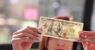 Kadın, ofisinde sahte para tespit etmek için 100 dolarlık banknotları kontrol ediyor. Bankada sahte elementler arayan kasiyer banknotları ışığa kadar tutuyor.