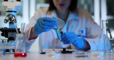 Bilim adamı şişenin içeriğini büyüteçle inceliyor. Odaklanmış laboratuar çalışanı, mavi ışık ağır çekimde steril laboratuvarda canlı sıvıyı inceliyor.