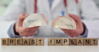 Doktor silikon implantları tutuyor ve boyut ve şekli kontrol ediyor. Minyatür tahta küpler masanın üzerinde durur ve BREAST İMPLANT ağır çekim ifadesini oluşturur