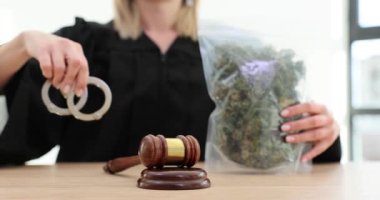 Yargıç masada oturmuş, kelepçe ve marihuana gösteriyor. Yetkili hakim, el konulan eşyalara dikkat çekiyor.