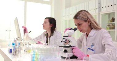 Tıp bilimcisi bir bilim laboratuvarında biyolojik örnekleri mikroskop altında inceliyor. Beyaz önlüklü güzel bir laboratuar mühendisi meslektaşıyla aşılar ve uyuşturucular üzerinde çalışıyor.