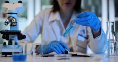 Laboratuvardaki bilim adamları deney tüpü ve mavi sıvıyla deney yapıyorlar. Kadın biyokimyacı test tüplerindeki sıvı örneklerini incelerken not alıyor.