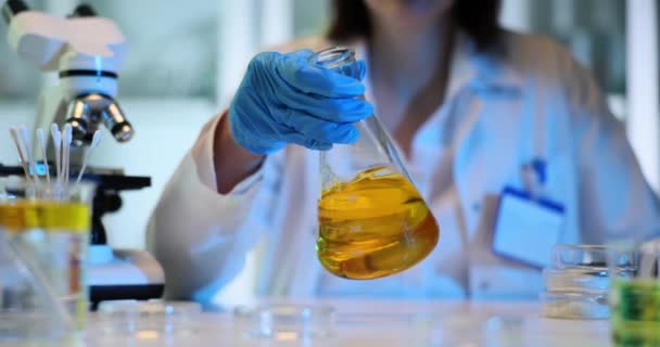 顕微鏡のクローズアップ前に黄色い液体でガラスフラスコを保持する科学者 液体を分析 試験する化学実験実験 — ストック動画