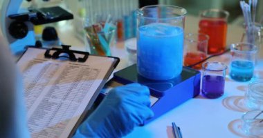 Bilim adamı manyetik dönüş kullanarak cam şişedeki mavi sıvıyı dikkatlice inceliyor. Laboratuvar asistanı laboratuarda tablet üzerine notlar alıyor