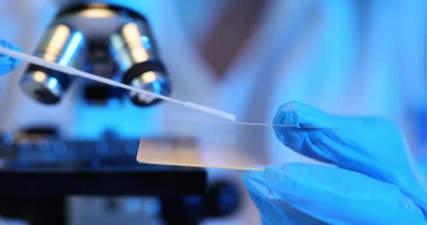 科学者はガラス顕微鏡の下でサンプルを分析する 生物学者の手袋の手はサンプルが付いているガラス スライドを握ります — ストック動画