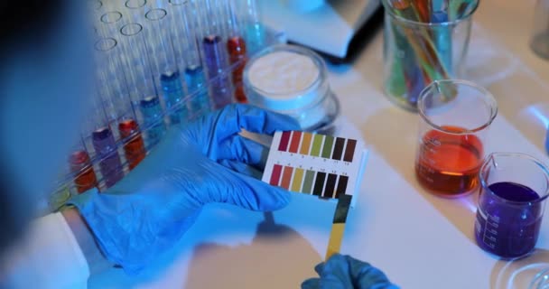 科学者はPhレベルをテストするために着色液のビーカーの隣にリットルマス紙ストリップを保持しています 手のクローズアップで テストを保持する科学者の化学者 — ストック動画