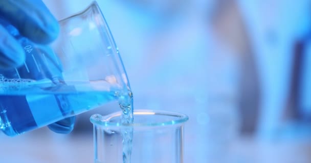 青い液体はガラスビーカーからフラスコに注がれます 塩基およびアルカリについての安定した反応を作成する青い解決が付いている試験管に化学薬品を注いで下さい — ストック動画