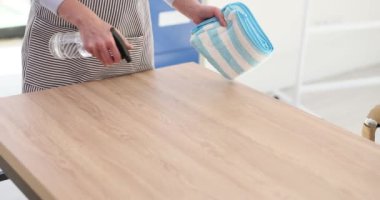 Kadın, toz ve lekeleri temizlemek için masayı bezle siler. Çalışkan kadın, deterjan şişesini ağır çekimde tutan mobilyaların yüzeyini spreyliyor.