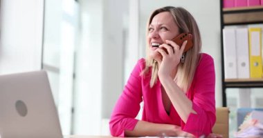 Kadın ofis çalışanı, iş yerinde dizüstü bilgisayarla oturarak akıllı telefondan konuşur. Sarışın kadın, dostça sohbete dalıp ağır çekimde keyif veriyor.