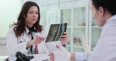 Kadın doktor, hastanın elinin yavaş çekimin röntgen görüntüsünü inceliyor. Profesyonel, klinikte eli bandajlı bir adama kalemle kırık olduğunu gösteriyor.