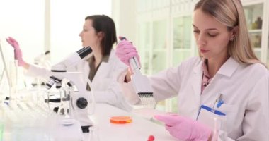 Eldivenli sarışın kadın daha fazla muayene için sıvı toplamak için cihaz kullanıyor. Üniformalı kadın kimyagerler laboratuvarda ağır çekimde çalışıyorlar.